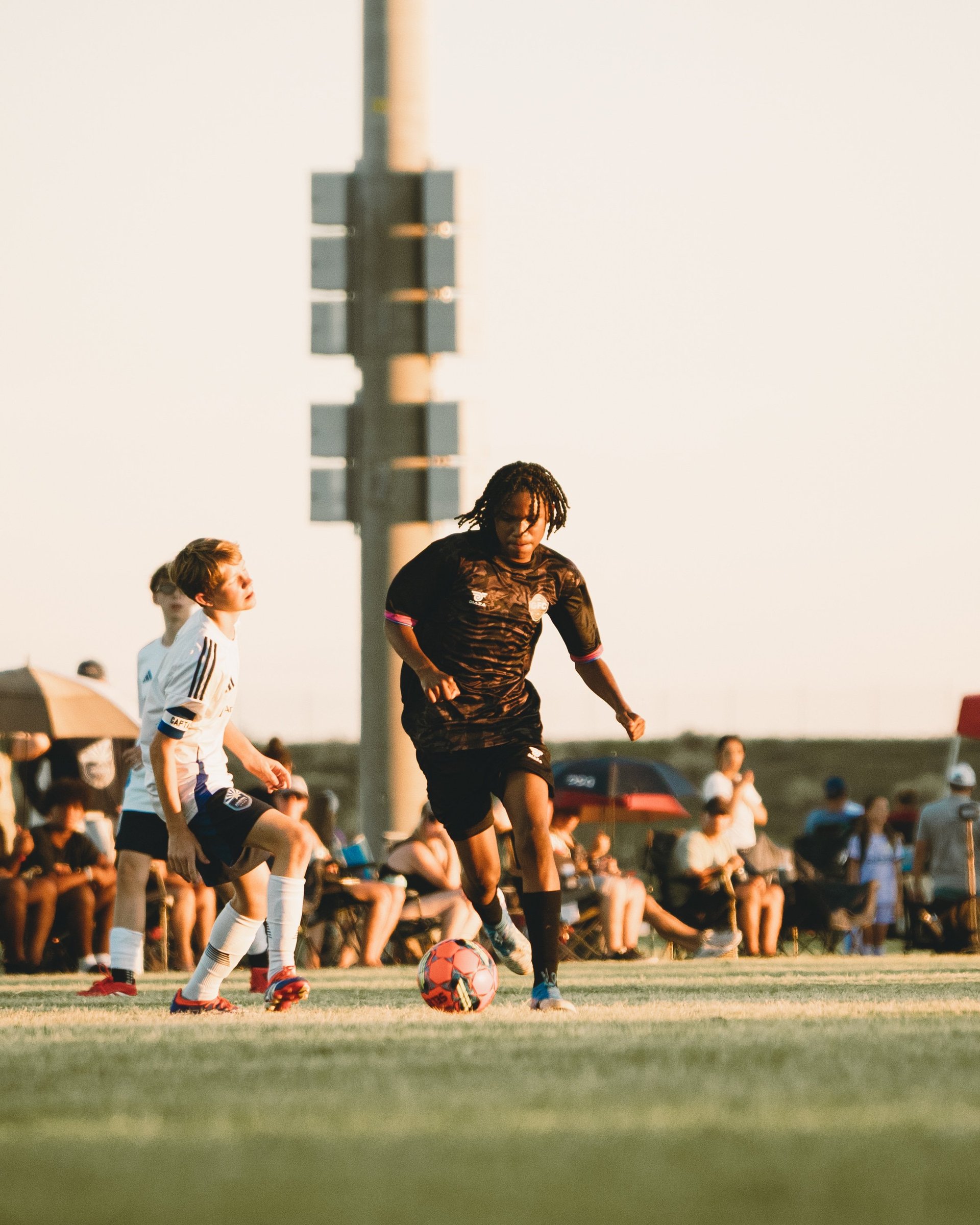 Young soccer players competing on a grass field during a youth soccer match with spectators and a tall stadium light tower in the background