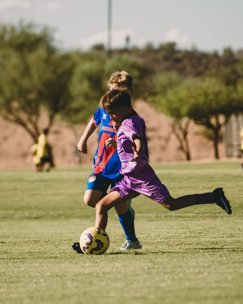 Young soccer players competing for the ball during a youth match on a grass field with trees and hills in the background