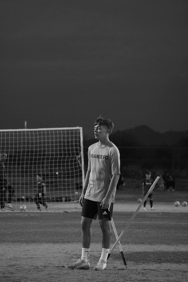 Black and white photo of a young soccer player standing on a field with a crutch, wearing a jersey with a soccer goal net visible behind him