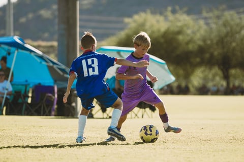 Two young soccer players competing for the ball during a youth match on a sandy field with blue umbrella and trees in background