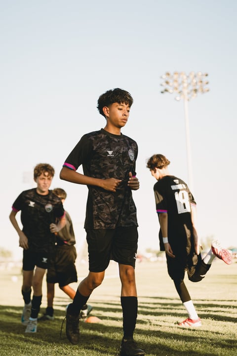 Three young soccer players in black uniforms running on a grass field during practice with stadium lights in the background