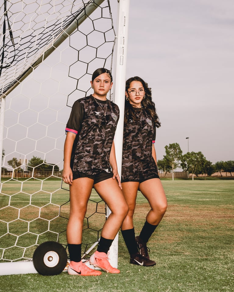 Two female soccer players in camo jerseys and black socks stand by a goal net with a soccer ball on a grass field