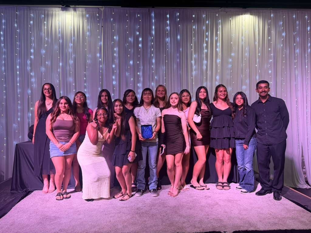 Large group of people in formal attire posing together in front of a shimmering purple curtain backdrop with dramatic lighting