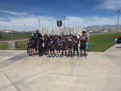 Youth soccer team in black uniforms posing together at sports complex with championship sign and fields in background