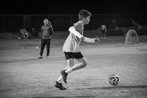 Young soccer player in white hoodie and shorts dribbling ball during evening practice on field