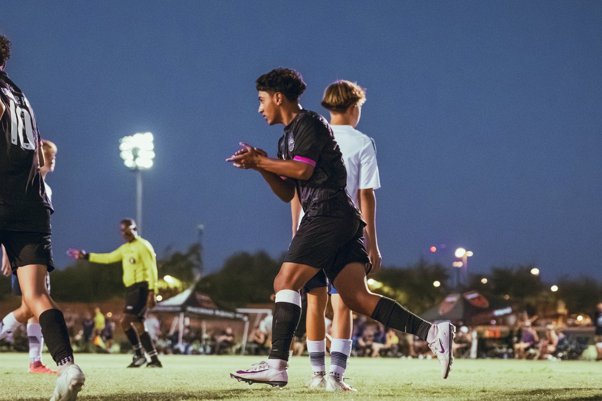 Female soccer players in black jerseys running with the ball during a night match under stadium lights with crowd in background