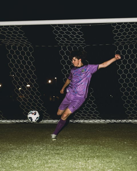 Soccer player in purple uniform performing a dynamic kick on a nighttime field in front of a goal net