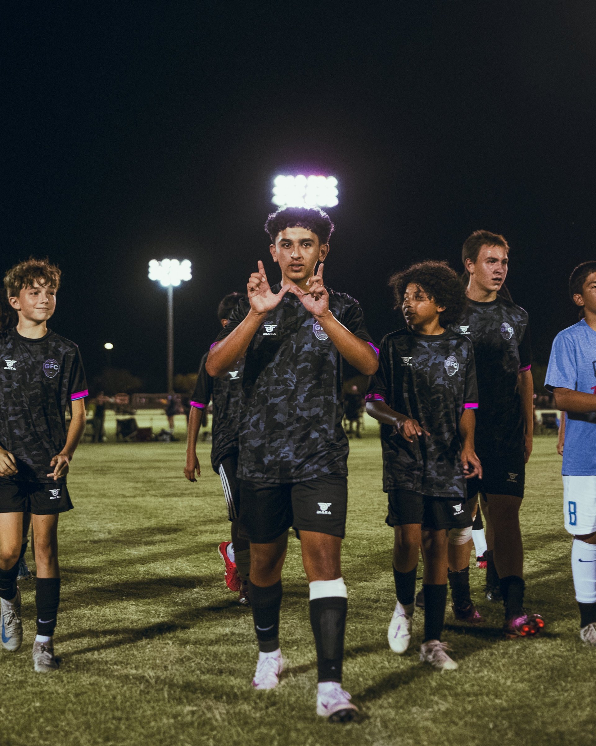 Youth soccer players in dark uniforms walk on a lit field at night, with a player in center gesturing and speaking to teammates
