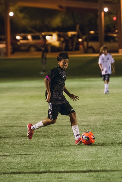 Young soccer player in black uniform dribbling orange ball on field at night with teammate in white visible in background