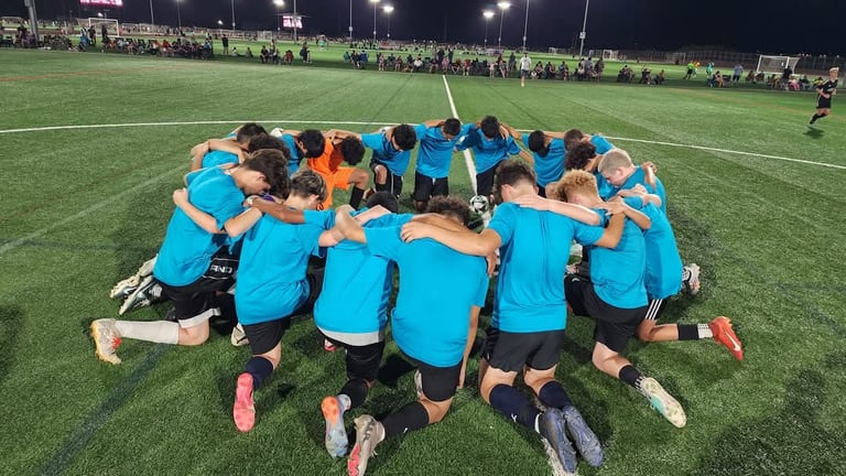 Youth soccer team in blue jerseys huddled together on field at night under stadium lights