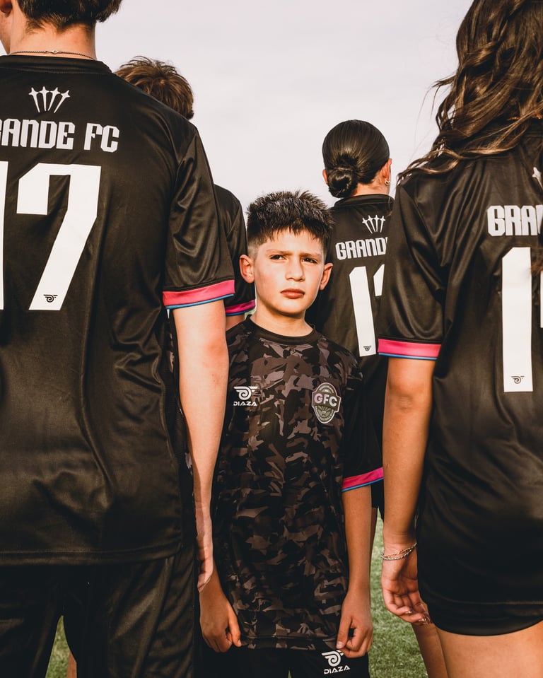 Young soccer player in black Grande FC jersey stands among teammates wearing matching uniforms with pink armbands on grass field