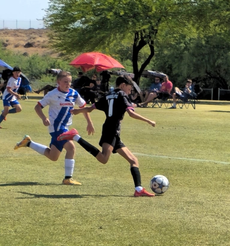 Young soccer players competing for the ball during a youth match on a grass field with spectators and red umbrella in background